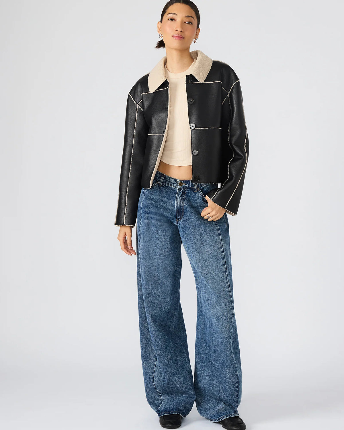Person wearing a black leather jacket with a white collar, beige top, and blue jeans on a white background